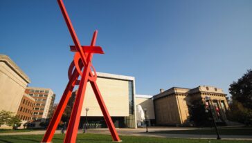 A large red sculpture stands tall over a green lawn. In the background, buildings silhouette a blue sky.