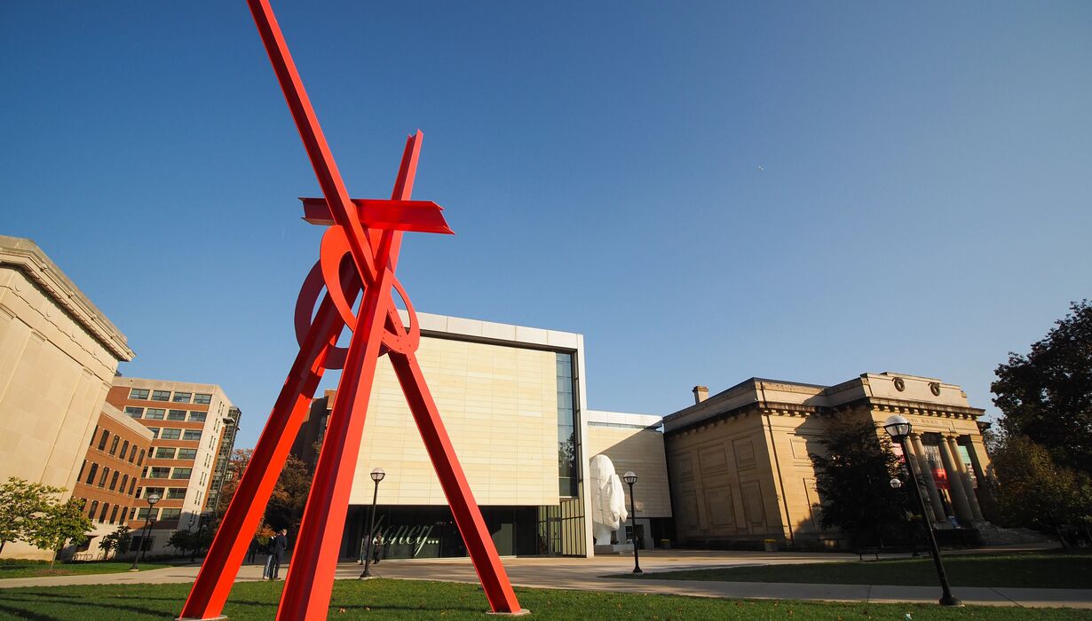 A large red sculpture stands tall over a green lawn. In the background, buildings silhouette a blue sky.