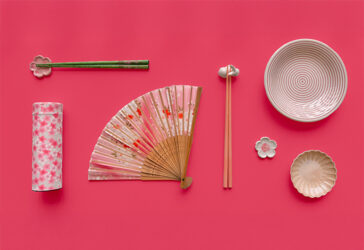 Flatlay of spring-inspired items arranged on a vibrant pink background. From left to right: a cylindrical tea canister decorated with pink cherry blossoms, a pair of green chopsticks resting on a pink flower-shaped chopstick holder, a pink and floral folding hand fan, a pair of pink chopsticks resting on a bunny-shaped chopstick holder, a cream-colored bowl with concentric pink rings, a beige dish with petal-like ridges, and a small white and pink flower-shaped chopstick holder.