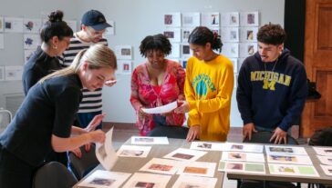 Five individuals collaborate around a table, examining and discussing various photographs and documents.