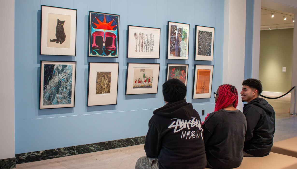 Three people sitting on a bench in an art gallery with vibrant red and blue walls and colorful artwork displayed.