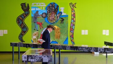 A person in a formal outfit examines an exhibit in an art gallery featuring vibrant murals with various figures and symbolic elements. Display tables with information are in the foreground.