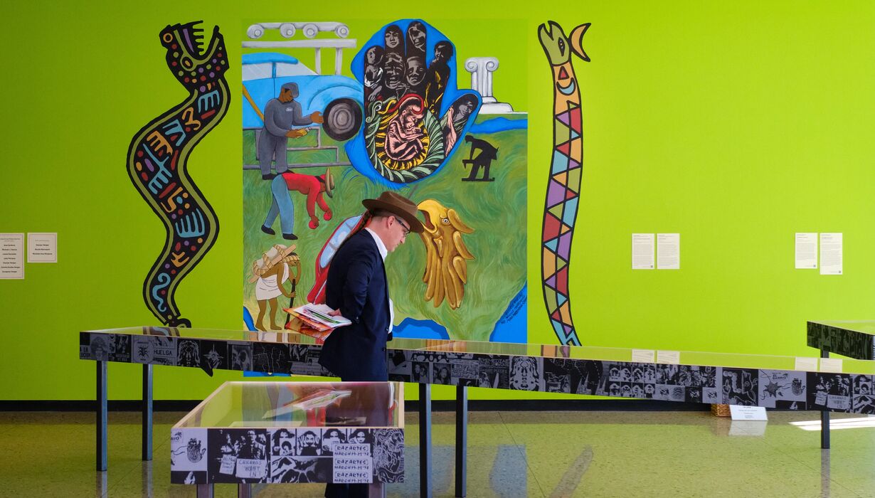 A person in a formal outfit examines an exhibit in an art gallery featuring vibrant murals with various figures and symbolic elements. Display tables with information are in the foreground.