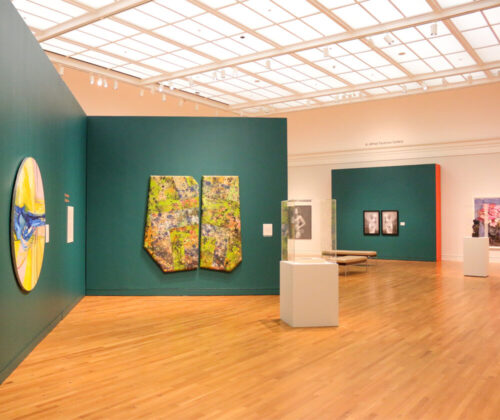 Art exhibition room featuring a large round canvas on the left, painted with bright colors and abstract patterns, and two rectangular canvases on the right showing detailed, colorful textures. The walls are green, and informational plaques are next to the artworks. A display case on the far right contains a sculpture.
