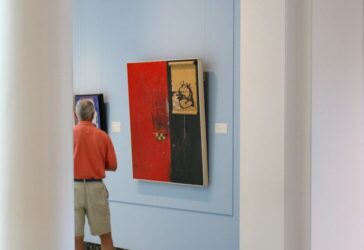 Person viewing abstract art pieces on display in a gallery, with one artwork prominently featuring red and black colors.