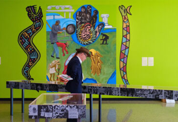A person in a formal outfit examines an exhibit in an art gallery featuring vibrant murals with various figures and symbolic elements. Display tables with information are in the foreground.