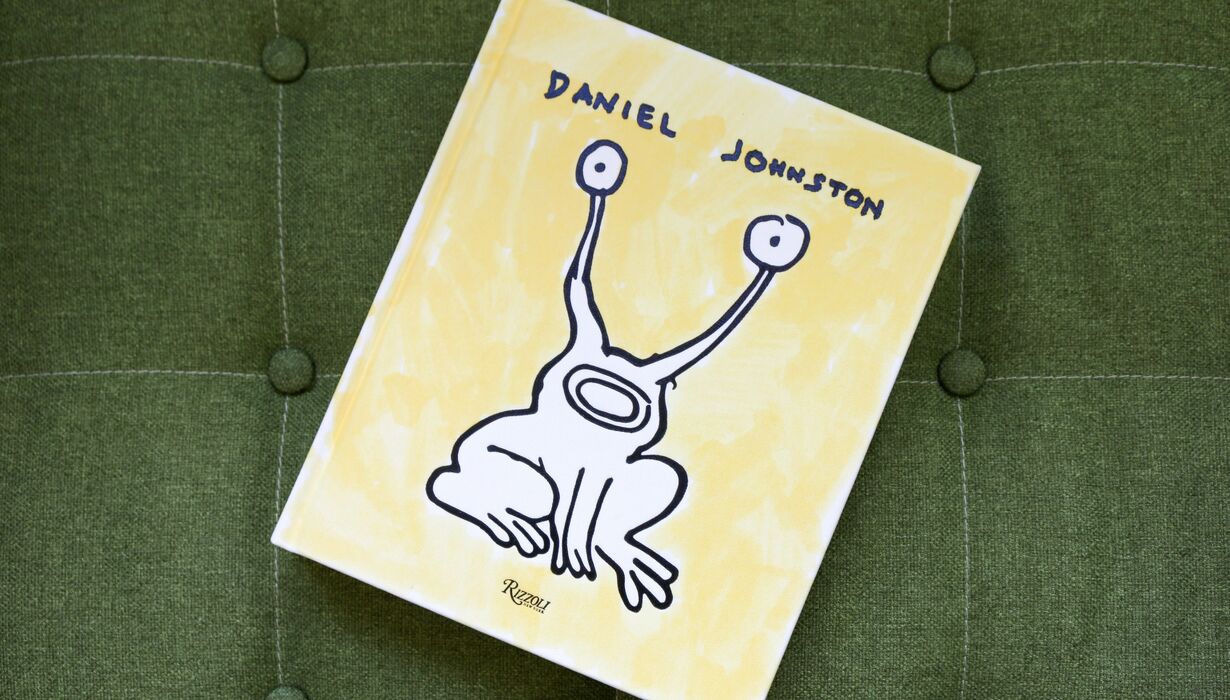 A book cover titled "Daniel Johnston" featuring a whimsical drawing of a frog-like creature on a yellow background, resting on a green cushioned surface with button details.