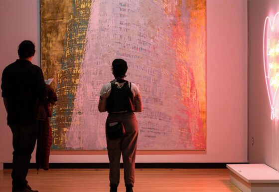 Image of two people backlight by a pink neon artwork looking at a large contemporary art object.