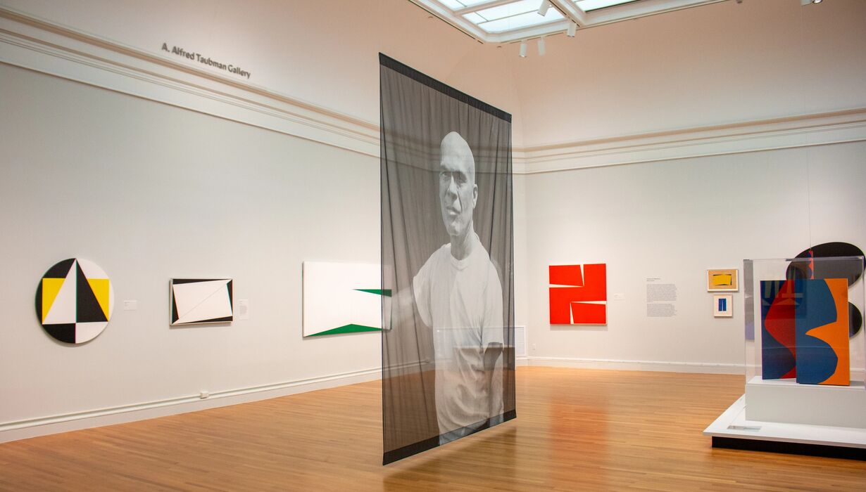 Art installation featuring a large, transparent panel with a monochrome photograph at an art gallery, surrounded by various abstract paintings.