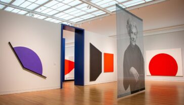 Interior of an art gallery showing abstract geometric artworks in vibrant colors displayed on white walls. Also featuring a large, transparent portrait of Carmen Herrera.