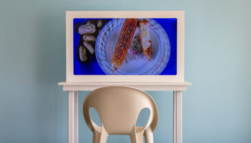 A modern chair is positioned in front of a small table displaying a video screen of a corn cob and sliced figs on a decorative plate. The setting is against a pale blue wall.