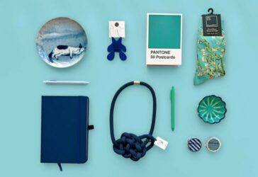A flat lay of art-inspired gift items arranged on a light teal background. Items include a ceramic dish with a illustration of a cat, a pair of abstract blue earrings, a box of Pantone 50 Postcards, Vincent van Gogh-inspired floral socks, a white pen, a green pen, a navy blue Moleskine notebook, a thick navy rope necklace with a decorative knot, a small scalloped ceramic dish, and two Washi tape rolls.