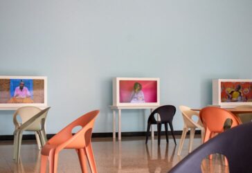 Two digital displays on white tables against a pale blue wall, showing videos of individuals. Both people are eating food and staring at the camera. Modern chairs are placed in front of the tables in a room with a minimalist style.