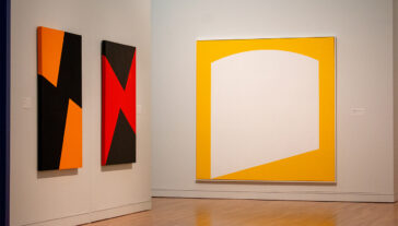 Three abstract paintings by Carmen Herrera, featuring sharp geometric patterns in black, orange, and white, displayed in an art gallery setting.