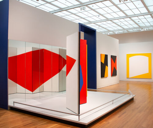 Installation view of a colorful abstract art piece featuring bold shapes and vibrant colors, displayed at a gallery with a blue wall background and natural lighting from a skylight.