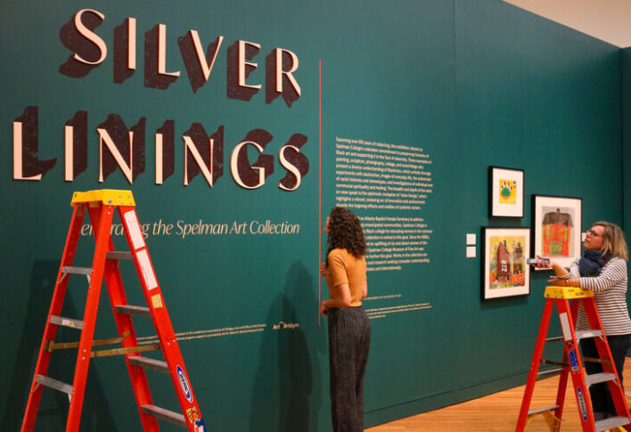 Two people view an exhibition titled 'Silver Linings: Celebrating 50 Years of the Spelman Art Collection' displayed on a teal wall. Two people observe the artwork and informational text.
