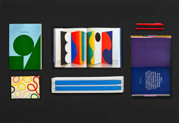 A flat lay of colorful stationery, art books, and candles on a black background. Items include a closed green-and-blue Essential Notebook, an open spread of the exhibition book “Both Sides of the Line,” a purple-and-blue patterned book with a quote printed inside, two red pens, a small notebook with colorful looping line designs, and a set of blue candles.