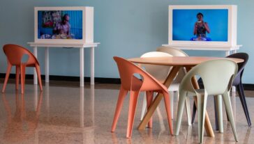 A modern beige chair facing a small white table with a digital screen displaying the text "Table Manners:" in a room with a light blue wall and beige floor.