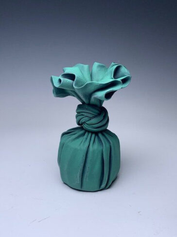 A trompe l’oeil ceramic sculpture that appears to be a green cloth wrapped around a small cup-shaped object and knotted. It is accompanied by an orange cloth and wooden box with a pink paper wrapping. The sculpture is painted on the lid of the box.