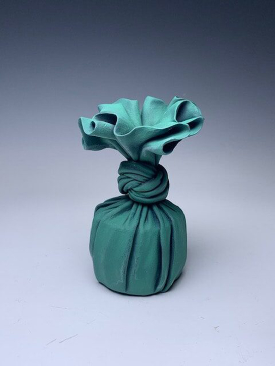 A trompe l’oeil ceramic sculpture that appears to be a green cloth wrapped around a small cup-shaped object and knotted. It is accompanied by an orange cloth and wooden box with a pink paper wrapping. The sculpture is painted on the lid of the box.