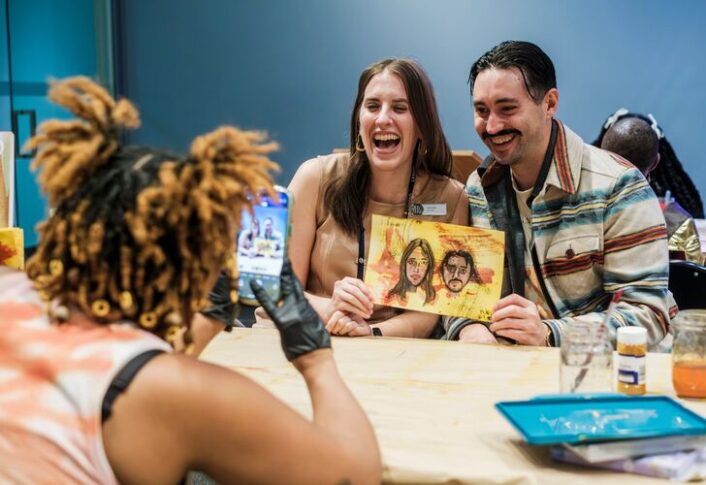 Person drawing a detailed portrait at a desk with watercolors and brushes nearby.
