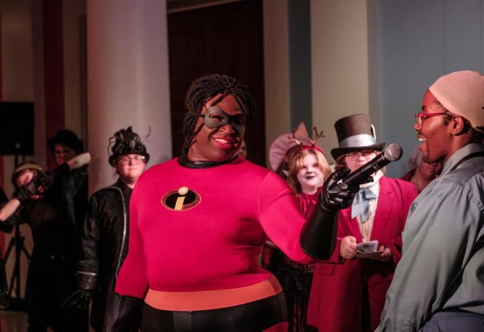 Person dressed as Mr. Incredible speaking into a microphone at an event with other attendees in various costumes in the background
