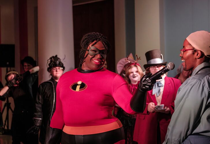 Person dressed as Mr. Incredible speaking into a microphone at an event with other attendees in various costumes in the background