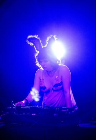 A DJ wearing rabbit ears and a pink top, performing at a music event with Pioneer DJ equipment, set against a backdrop of a spacious, elegant venue with columns.