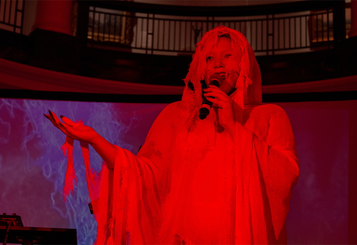A person performs on stage dressed in a white, ghostly costume with flowing fabric, under red lighting.