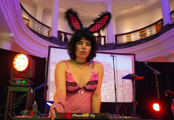 A DJ wearing rabbit ears and a pink top, performing at a music event with Pioneer DJ equipment, set against a backdrop of a spacious, elegant venue with columns.
