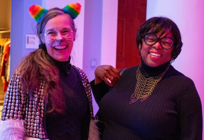 Two individuals smiling at a social event, one wearing a multicolored headband and the other sporting a stylish necklace and glasses.