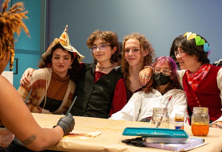 Group of young people wearing casual and costume attire, including hats and a mask, gathered around a table with drinks and a book, smiling andgeeting their portraits drawn, interacting with each other.