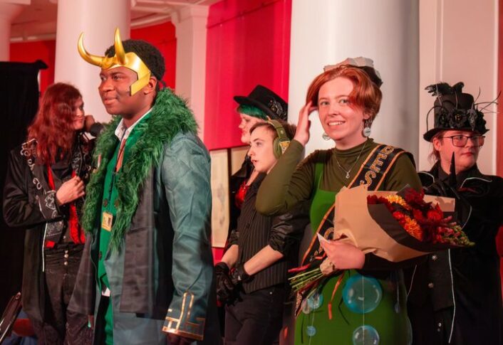 A group of people in costume at a themed event, featuring individuals dressed as a character with a horned helmet and another with a green outfit adorned with bubbles, among others in various imaginative outfits.