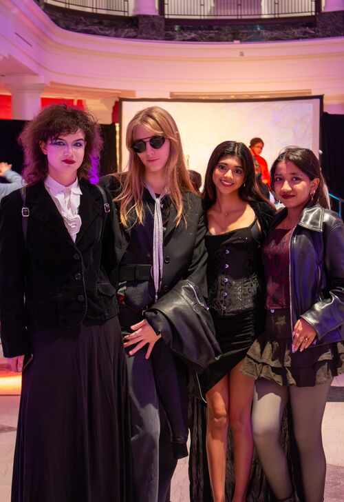 Four individuals in Gothic-inspired outfits posing together at an event, featuring varied styles including ruffled shirts, corsets, sunglasses, and layered jewelry.