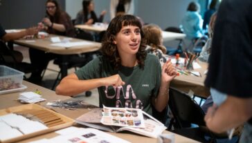 Person wearing shirt with UMMA logo sits and talks with someone at table with art supplies