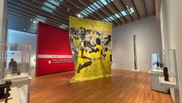 A large floor-to-ceiling textile hangs from the ceiling of an art gallery. The textile is yellow, grey, and brown and depicts individuals styling others' hair.