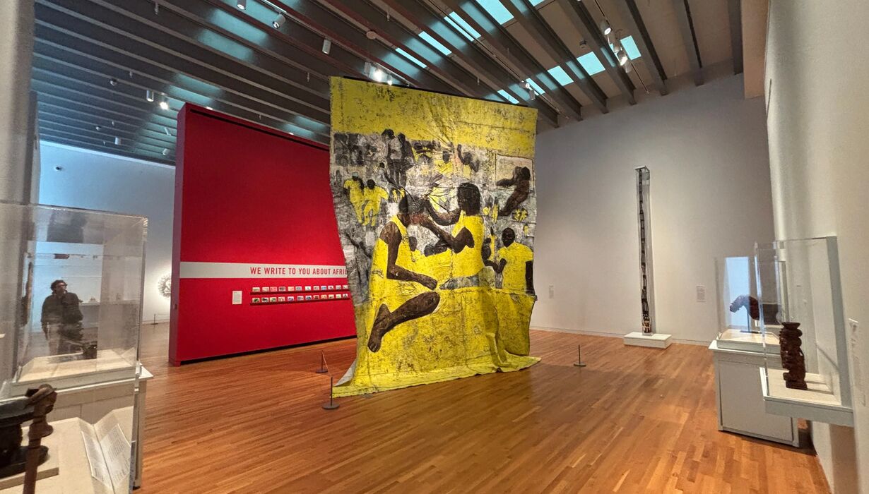 A large floor-to-ceiling textile hangs from the ceiling of an art gallery. The textile is yellow, grey, and brown and depicts individuals styling others' hair.