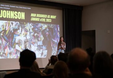 Students stand on stage in front of a large projection screen with presentation behind them.