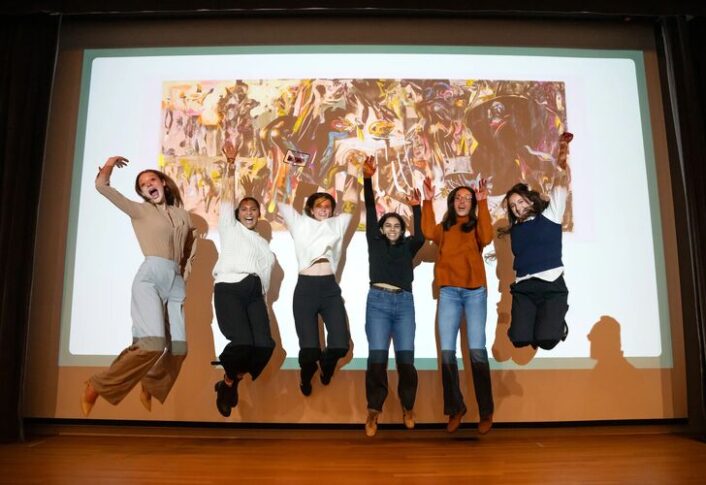 Students jump on stage in front of a large projection screen with microphone. The screen reads "ART TANK"