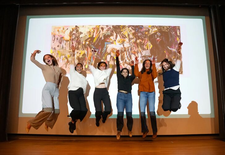 Students jump on stage in front of a large projection screen with microphone. The screen reads "ART TANK"