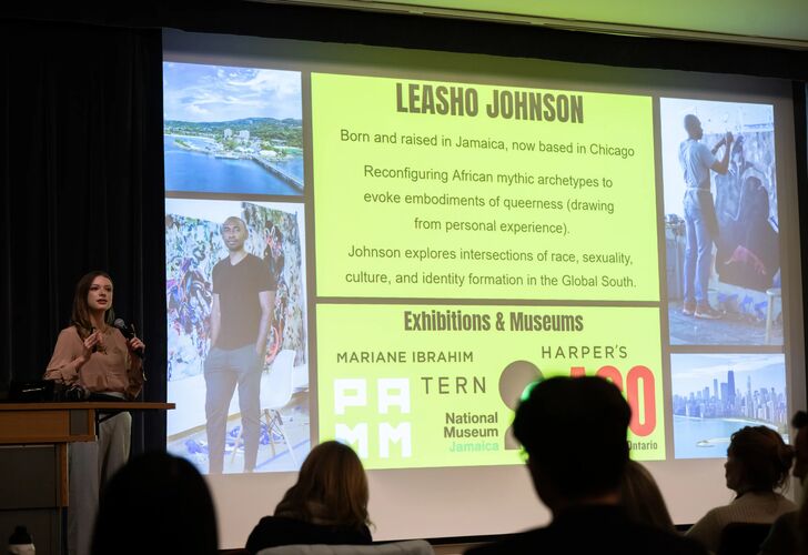 Students stand on stage in front of a large projection screen with presentation behind them.