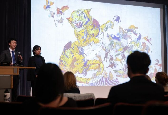 Students stand on stage in front of a large projection screen with presentation behind them.