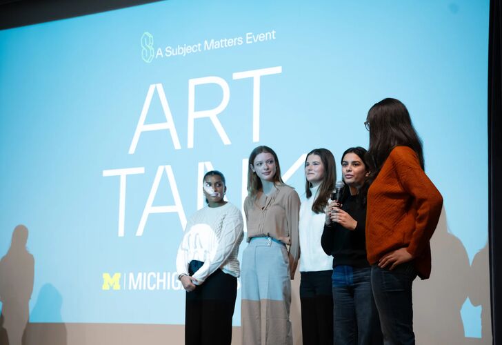 Students stand on stage in front of a large projection screen with microphone. The screen reads "ART TANK"