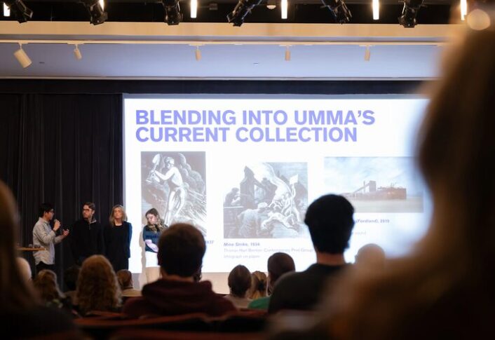 Students stand on stage in front of a large projection screen with presentation behind them.
