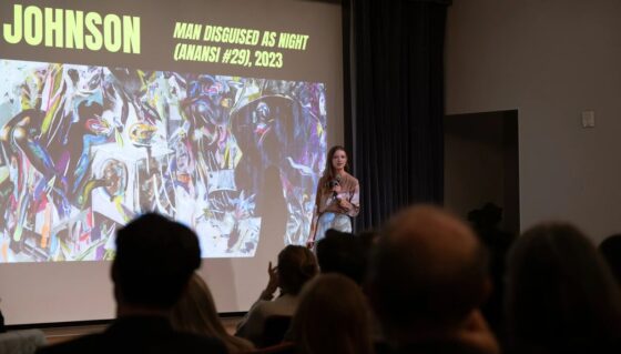 People stand on stage in front of a large projection screen with microphone.