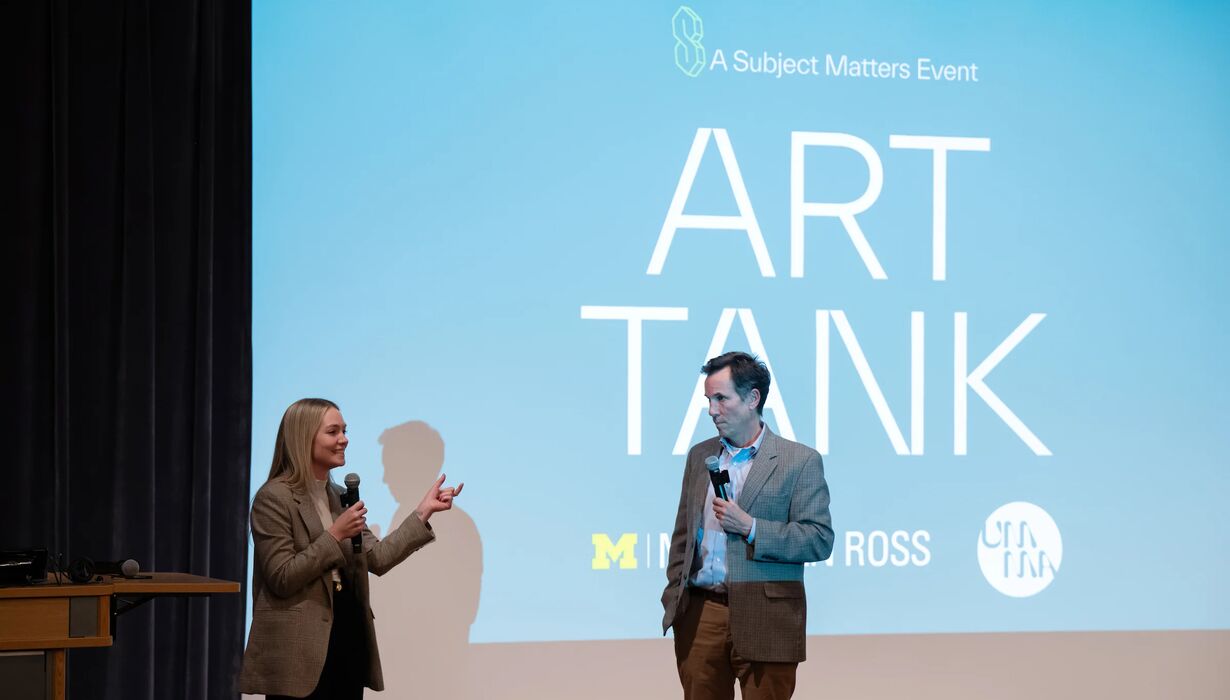 People stand on stage in front of a large projection screen with microphone. The screen reads "ART TANK"