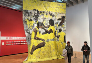 A large floor-to-ceiling textile hangs from the ceiling of an art gallery. The textile is yellow, grey, and brown and depicts individuals styling others' hair.