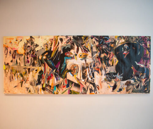 This image shows a modern art gallery with wooden floors and glass railings. Several artworks are displayed on the walls, including a large colorful abstract painting and smaller framed pieces. Bright natural light streams in from a window on the right.