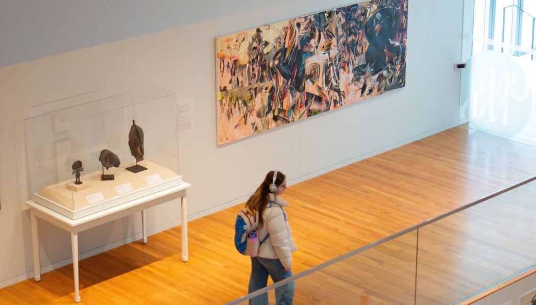 This image shows a modern art gallery with wooden floors and glass railings. Several artworks are displayed on the walls, including a large colorful abstract painting and smaller framed pieces. Bright natural light streams in from a window on the right.