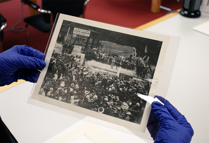 Photo of a person with blue gloves on holding a photo titled "Laying of the Corner Stone of the Alumni Memorial Building, June 17, 1908 (Photo by Lyndon)/HS671" from the image archives at the Bentley Historical Library.